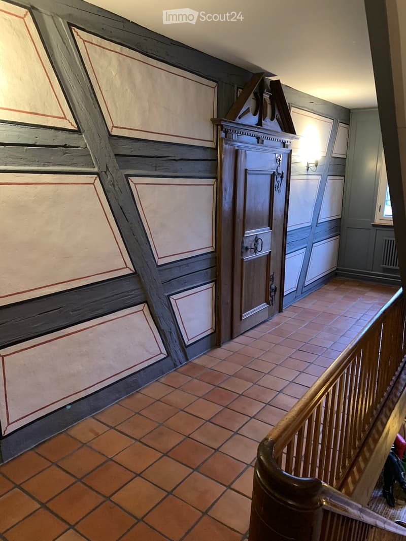 Staircase with wooden handrail, terracotta tiles, and painted walls with geometric patterns. A closed door is visible at the end of the hallway.