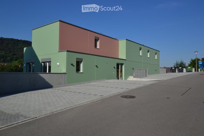 Modern two-story building with green and red exterior walls, multiple windows, and a raised ground floor entrance area.
