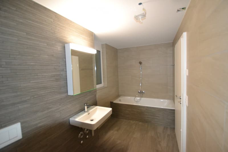 Modern bathroom with a white sink, bathtub, and illuminated mirror. The walls are tiled in a neutral color scheme, and the floor is wooden.