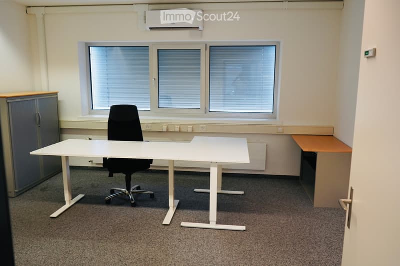 Office space with a white desk, black office chair, and a small cabinet. Large windows with blinds and an air conditioning unit are visible.