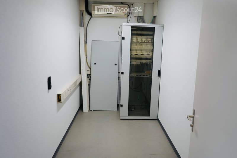 Empty room with white walls and a tiled floor, containing a server rack, an air conditioning unit, and electrical panels.