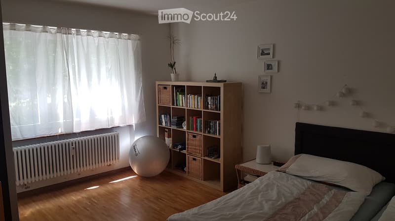 Cozy bedroom with wooden floor, large window with white curtains, bed with pillows and blanket, bookshelf filled with books and baskets, round exercise ball, small table with lamp, and decorative string lights on the wall.