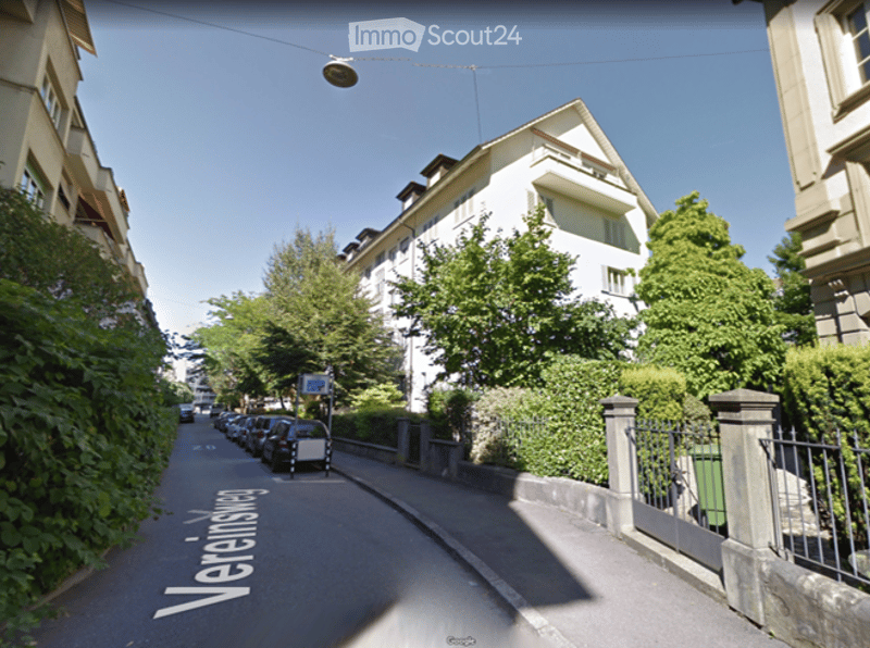 A quiet residential street lined with parked cars and multi-story apartment buildings, surrounded by lush green trees and shrubs.