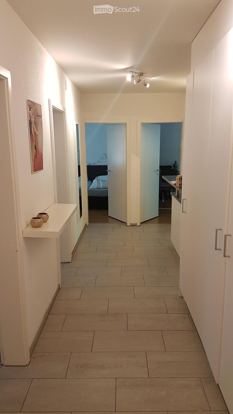 A narrow hallway with tiled flooring, white walls, and recessed lighting. Two doors lead to a bedroom and a kitchen area, with a small shelf holding decorative items on the left side.