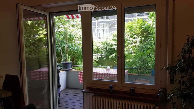 Balcony with a checkered tablecloth-covered table, chairs, and potted plants, overlooking a lush green garden.