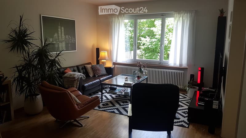 Modern living room with large windows, leather sofa, glass coffee table, armchair, table lamp, and decorative plants.