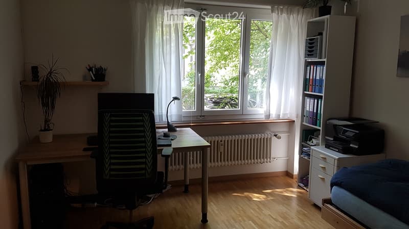 Well-lit room with wooden flooring, large window with white curtains, desk with chair, screen, lamp, shelf with binders, printer, and a small bed.