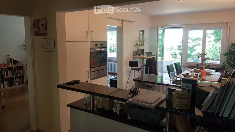 Bright, modern kitchen with white cabinets, stainless steel oven, and a dining area with a table and chairs. The counter holds various items including jars, cups, and a vase with flowers.