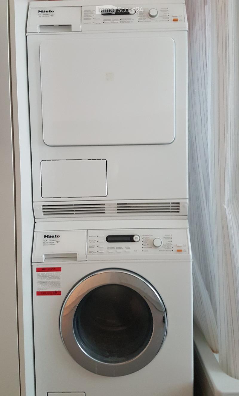 White Miele washing machine with control panel and dials, placed against a light-colored wall.