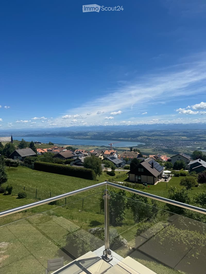 Balcony with a modern glass railing, overlooking a scenic landscape with a lake, mountains, and houses.