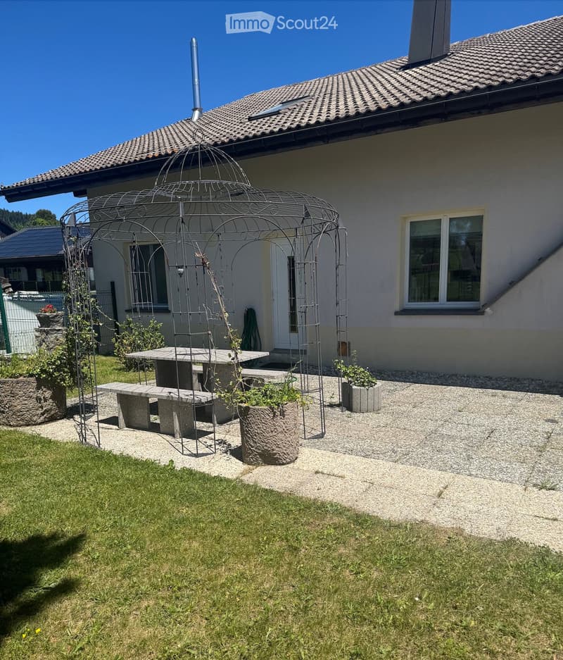 Garden area with a stone table, benches, a decorative metal arch, and potted plants near a house.