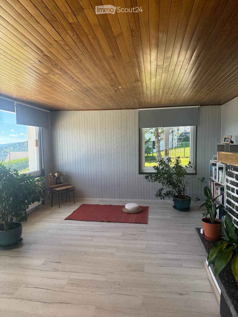 Bright, minimalist room with wooden floors and a slanted wooden ceiling. Features large windows with roller shades, a small table with a lamp, a bookshelf, potted plants, and a red meditation mat with cushions.