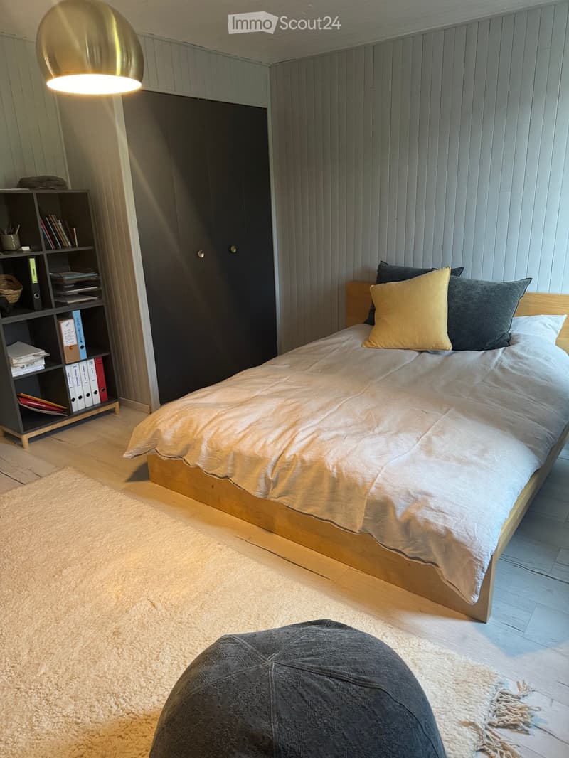 Modern bedroom with a wooden bed, beige bedding, grey and yellow pillows, a round lamp, a bookshelf, and a grey pouf on a light-colored carpet.