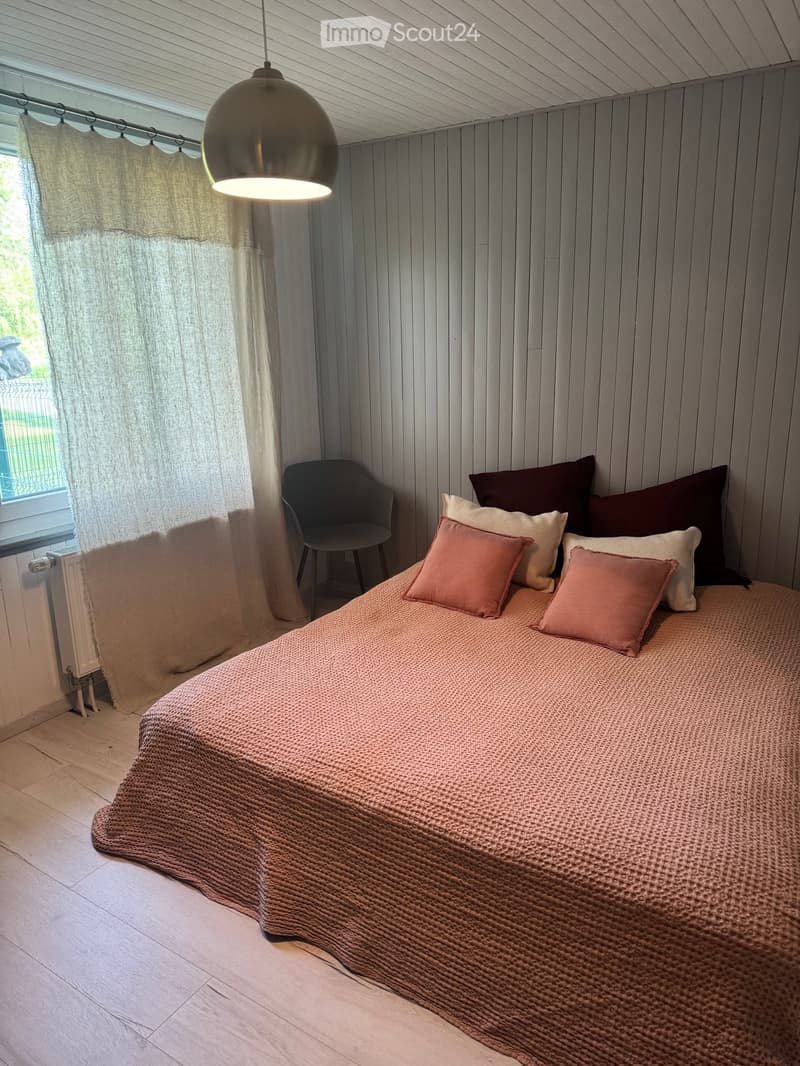 A cozy bedroom with a neatly made bed, pillows, a chair, and a hanging pendant light. The walls are paneled in light wood, and the room has a large window with curtains.