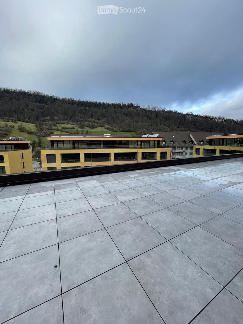 Roof terrace with large, light-colored tiles and a modern, sleek design. The terrace overlooks a modern building complex with yellow and orange facades and large glass windows, set against a backdrop of green hills and a partly cloudy sky.