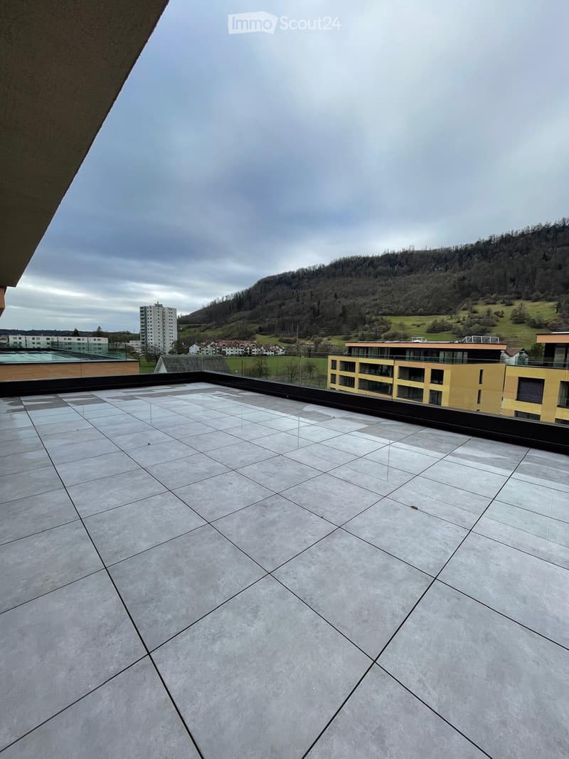 Roof terrace with large, light grey tiles, glass railing, and a view of a hilly landscape with buildings and greenery.