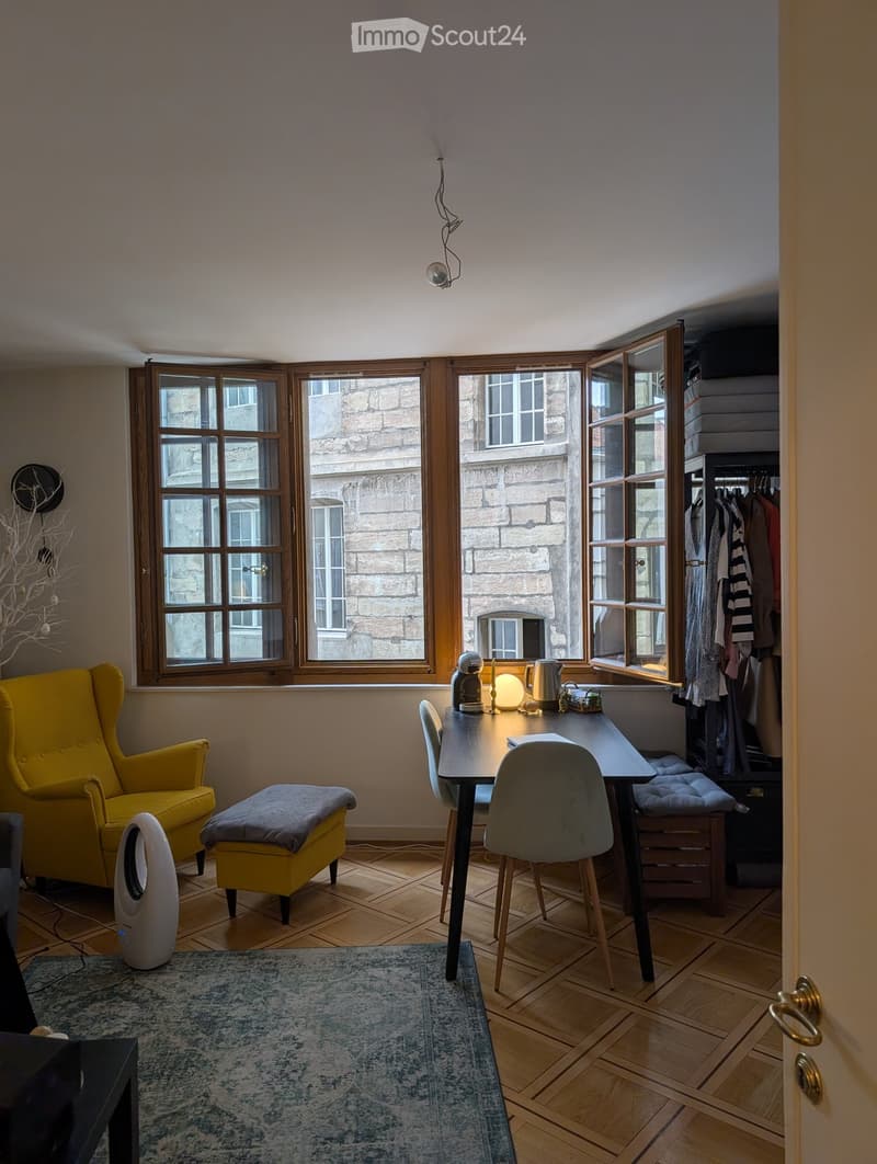 A cozy, well-lit room with wooden floors, large windows, a yellow armchair, a small table with chairs, a stool, a lamp, a teapot, a mirror, and a wardrobe with hanging clothes.