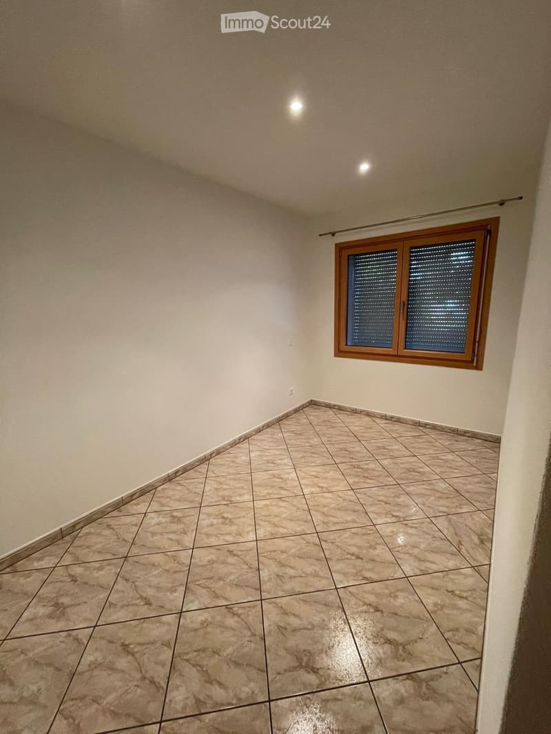 Empty room with beige tiled flooring and white walls, featuring a window with blinds and a ceiling light fixture.