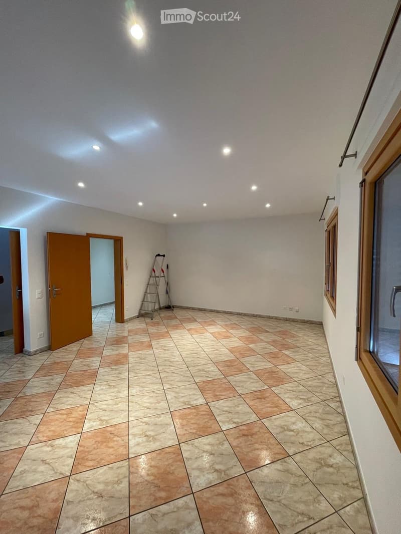 Empty room with tiled floor, white walls, and recessed lighting. Features a wooden door, a window with wooden frame, and a ladder in the corner.