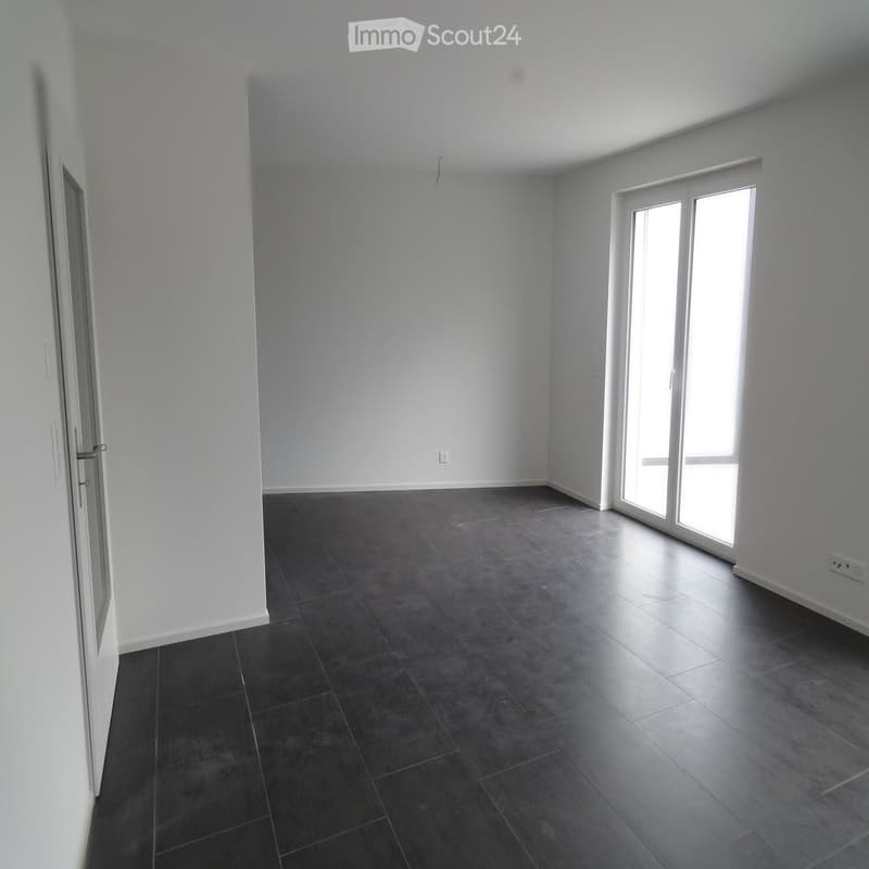 Empty room with dark grey tiled floor, white walls, and large windows letting in natural light.