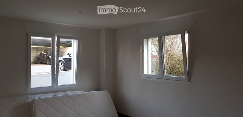 Empty bedroom with a single bed, large windows, and a sliding door leading to a garage with parked cars.