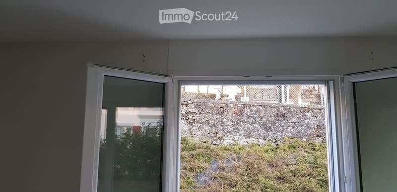 Empty room with large windows showing a view of a stone wall and grassy area outside.