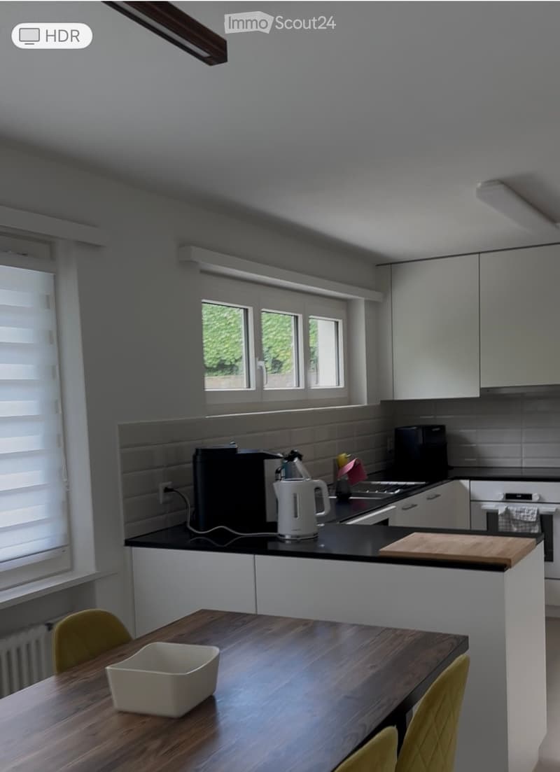 Modern kitchen with white cabinets, black countertops, and a wooden dining table with yellow chairs. Appliances include a microwave, kettle, and oven.