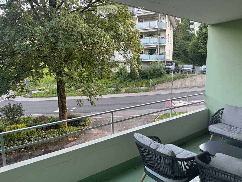 Balcony with two wicker chairs, a small table, and a railing overlooking a street with trees and parked cars.