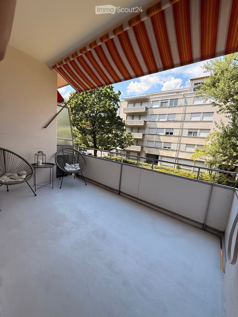 Balcony with a striped awning, two modern chairs, a small table with a candle, and a view of a multi-story building and trees.