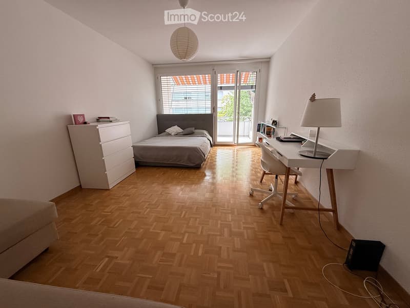 Bright bedroom with wooden flooring, a double bed with grey bedding, a nightstand, a desk with a chair, and a hanging lamp.