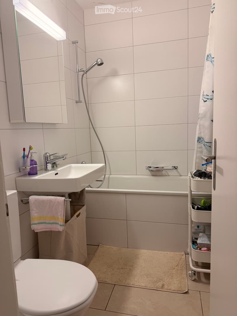 Modern bathroom with white tiles, a sink, toilet, bathtub, and a towel on a rack. A small cart holds various items.
