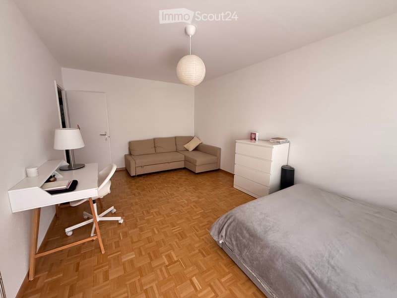 A furnished room with a bed, sofa, desk, chair, and a lamp. The room features wooden flooring and white walls.