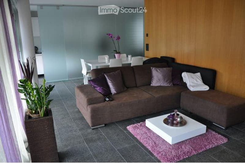 Modern living room with a grey sectional sofa, white coffee table, dining area with white table and chairs, and decorative plants.