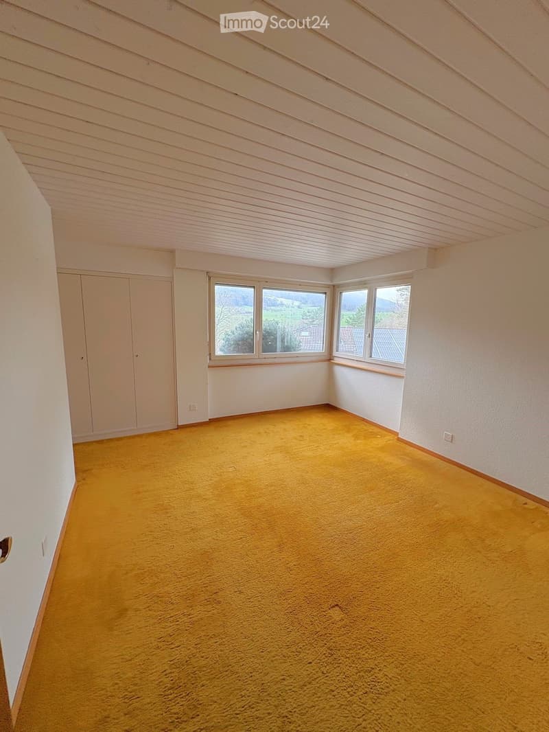 Empty room with yellow carpet, white walls, and large windows offering a view of the landscape.