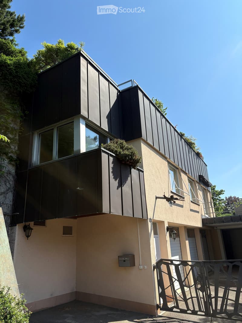 Modern two-story building with a combination of dark metal siding and light beige walls, featuring large windows, a small balcony with greenery, and a gated entrance.