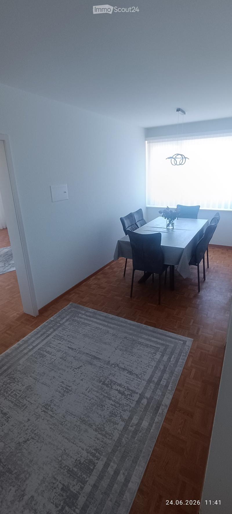 Dining room with wooden flooring, a large dining table surrounded by six chairs, a table runner, and a vase with flowers on the table. A patterned rug is placed on the floor near the table.