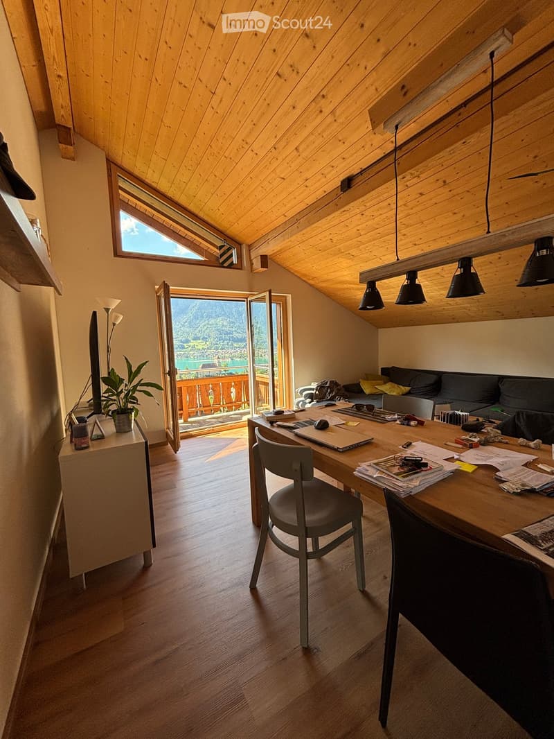 Bright, wooden-floored room with a slanted wooden ceiling, large windows, and a view of a lake and mountains. Features a sofa, desk with laptop, chair, and a potted plant.