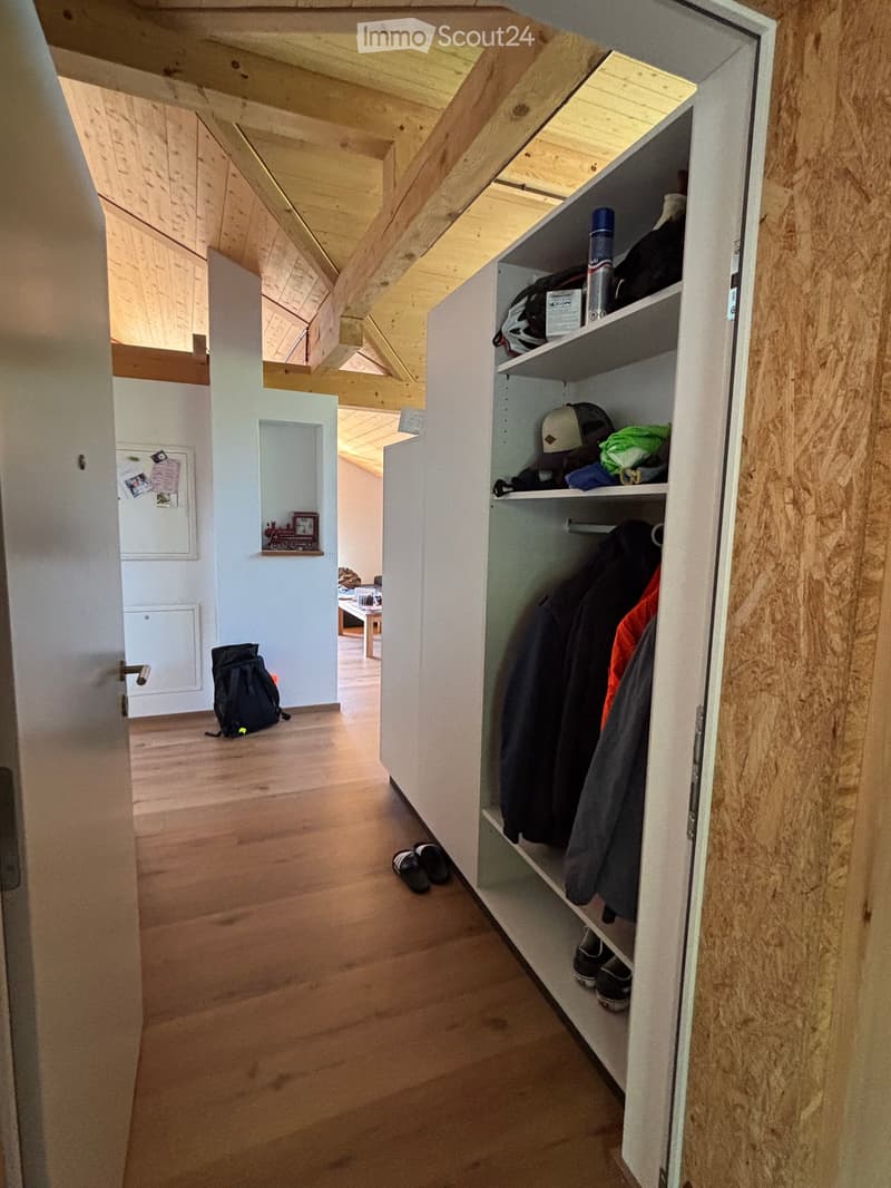 A wooden-floored hallway with exposed wooden beams on the ceiling, featuring a built-in closet with hanging clothes and shelves holding various items, including shoes and a backpack.