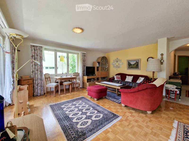 Bright living room with wooden flooring, large windows, a red sofa, coffee table, ottoman, dining area with chairs and table, television, and decorative items.