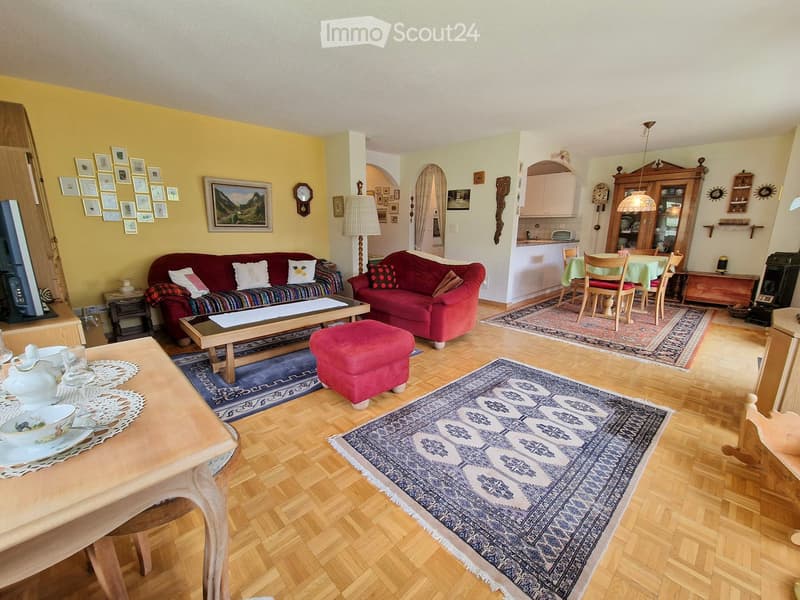 Bright living room with wooden flooring, two red sofas, coffee table, ottoman, dining area with table and chairs, and a kitchen area in the background.