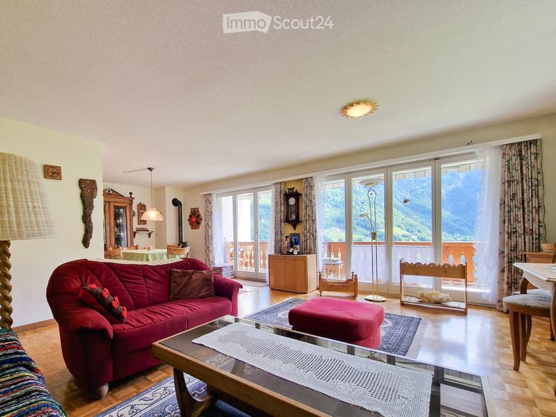 Bright living room with wooden flooring, large windows offering a scenic mountain view, red sofa, coffee table, ottoman, dining area with table and chairs, and decorative cabinet.