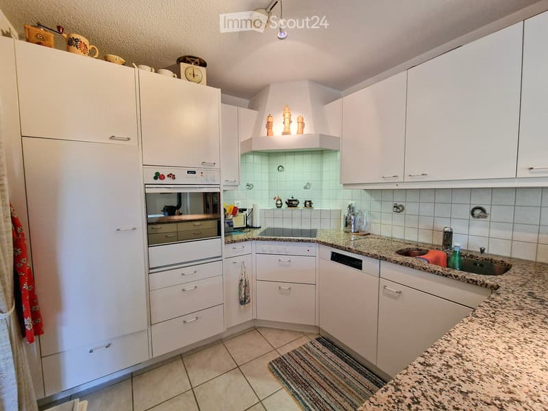 A modern kitchen with white cabinets, granite countertops, and stainless steel appliances including a refrigerator, oven, and dishwasher. The kitchen features a tiled backsplash and a small rug on the floor.