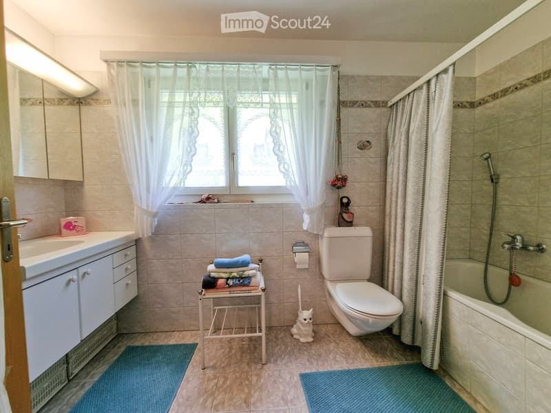 Bathroom with beige tiles, a sink with a cabinet, a toilet, a bathtub with a shower, a towel rack with folded towels, a small cat figurine, and a blue bathmat on the floor.