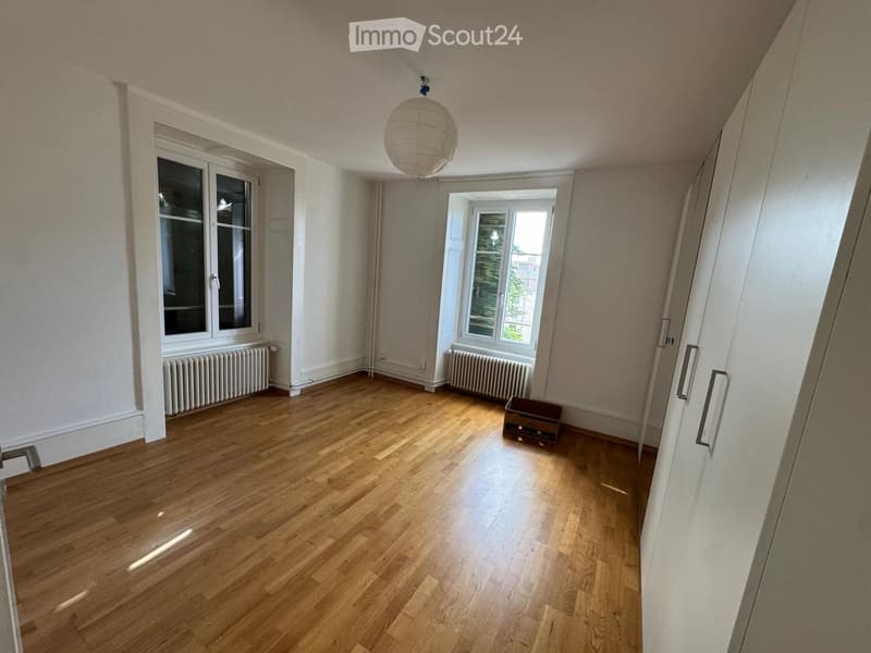 Empty room with wooden flooring, white walls, large windows, radiators, and a hanging light fixture.
