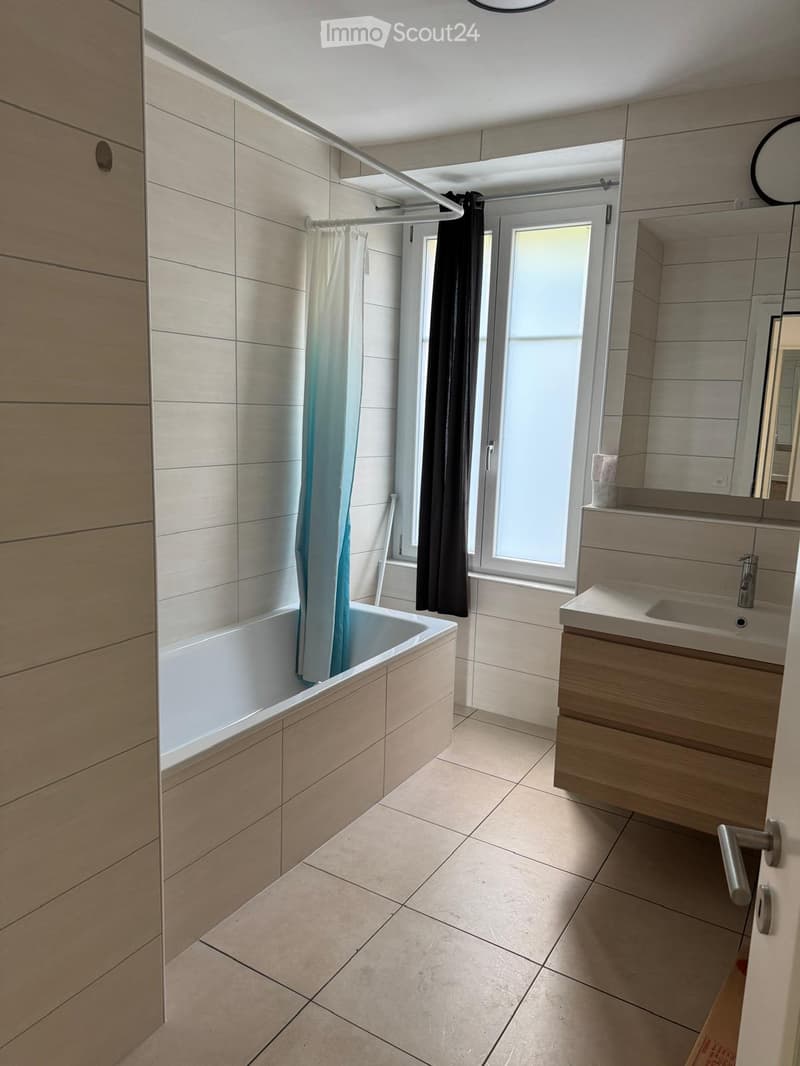 Modern bathroom with a bathtub, sink, and mirror. Light-colored tiles and a window with a curtain provide natural light.