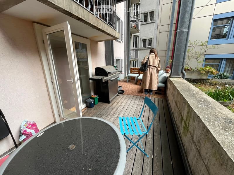 Balcony with wooden decking, round table, folding chair, grill, small bench, and potted plants.