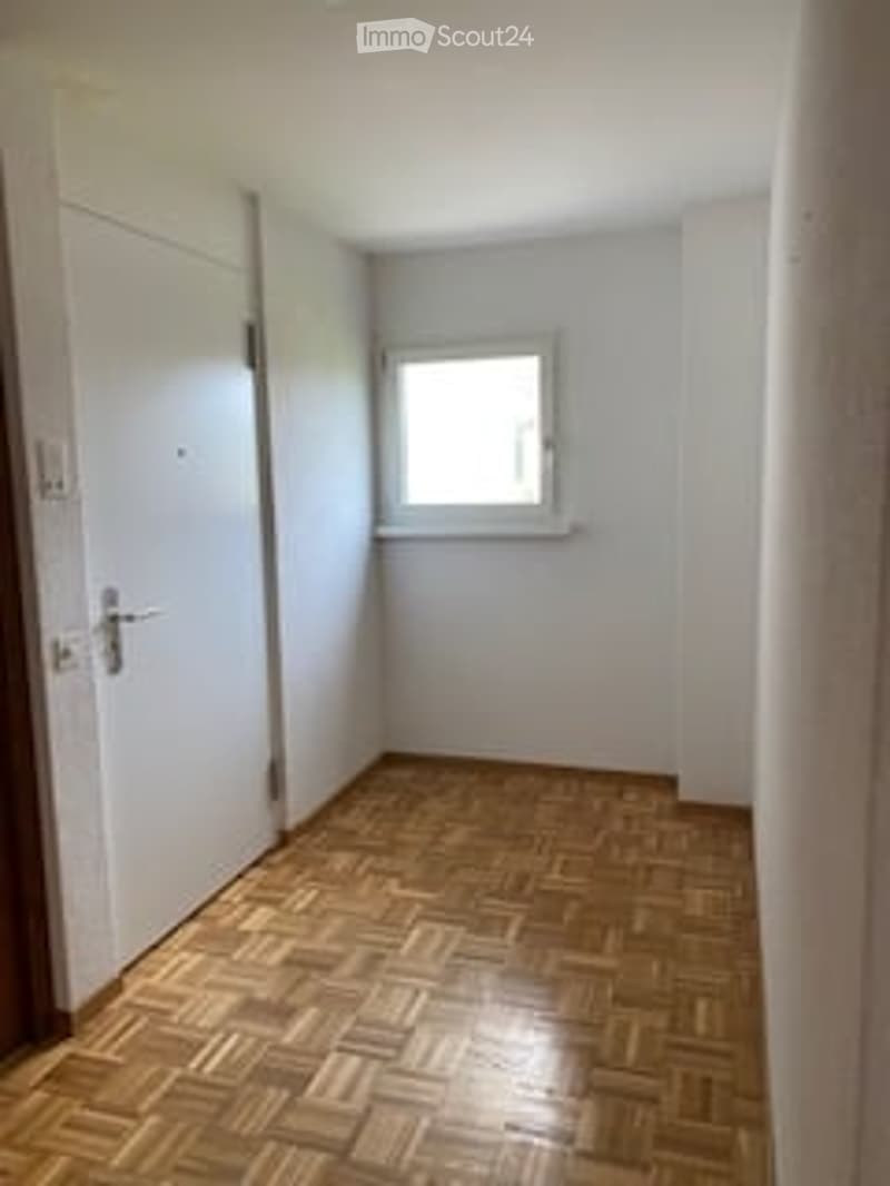 Empty room with wooden parquet flooring, white walls, and a single window with white frame. No furniture or equipment present.