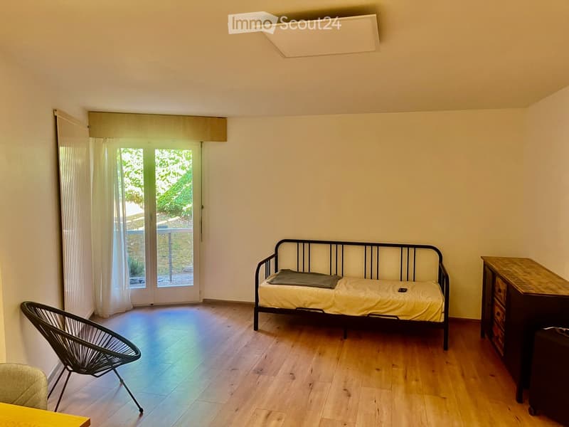A small, furnished room with a wooden floor, a sofa, a chair, a small table, and a cabinet. The room has large windows with curtains and a door leading to a balcony.