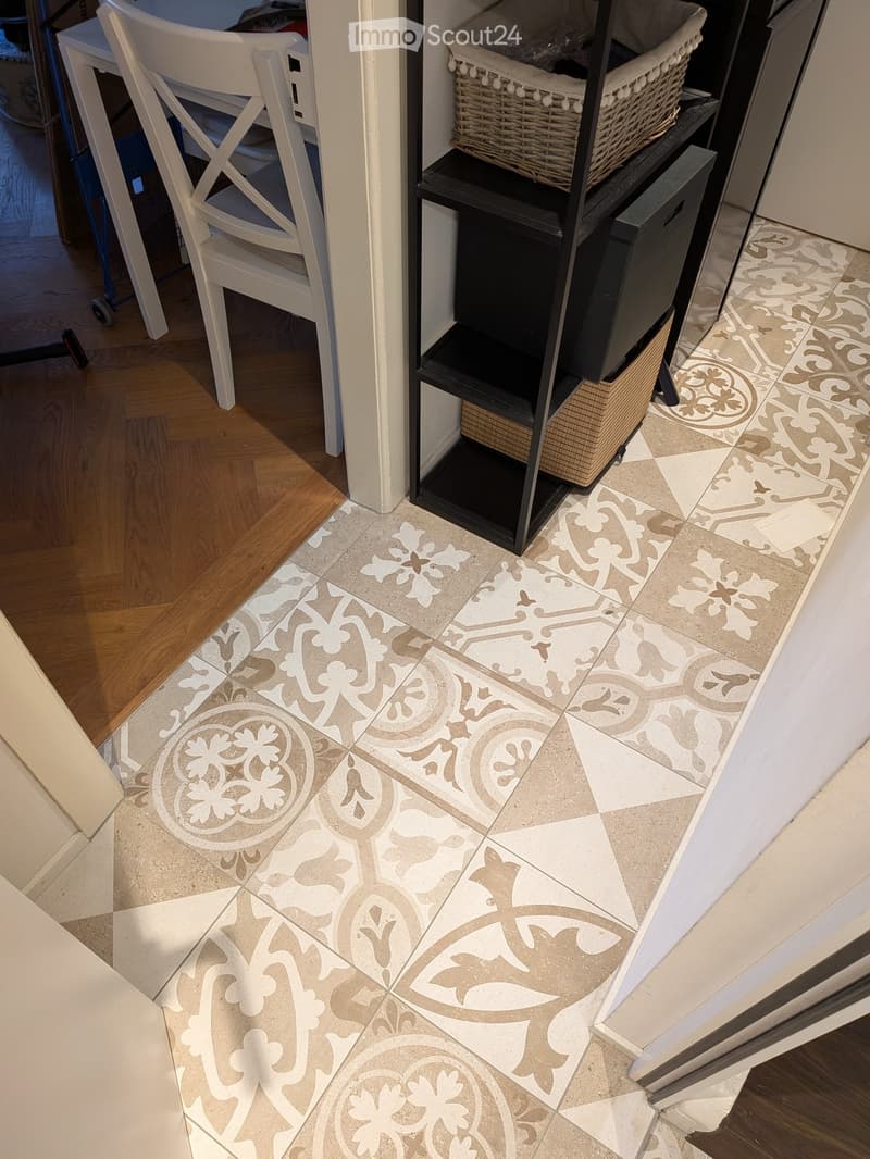 Hallway with decorative tile floor featuring floral and leaf patterns in beige and grey tones, adjacent to wooden flooring and white furniture.