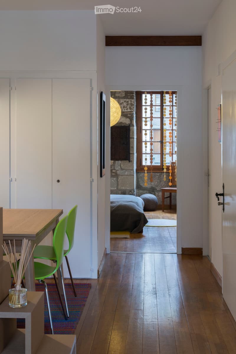 A modern hallway with wooden flooring, white walls, and a view into a bedroom area with a bed, chair, and decorative elements.